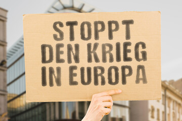 The phrase " Stop the war in Europe " is on a banner in men's hands with blurred background. Support. Community. Friendship. Dialogue. Harmony. Reconciliation. Resolution. Peacemaker. Antiwar. Pacifis
