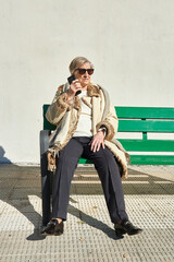 an eighty-year-old woman sunbathes in sunglasses sitting on a green wooden bench in front of a concrete wall while talking on the cellular phone