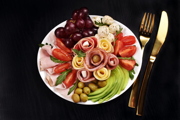 Top view of a platter with assortment of sliced ham and cheese with vegetables and grape on black...