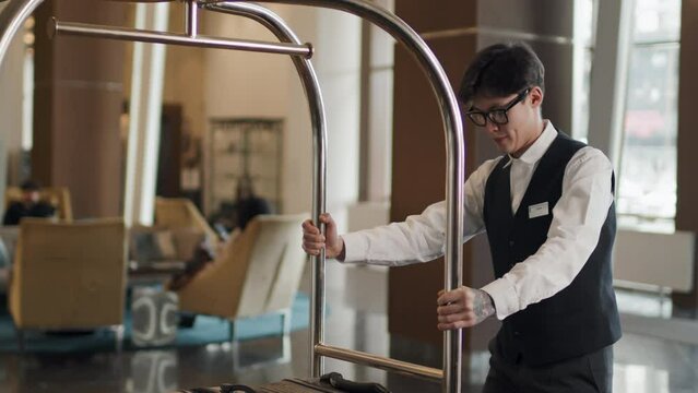 Slowmo of young Asian man working as hotel porter or bellhop in modern hotel using luggage trolley to transport suitcases