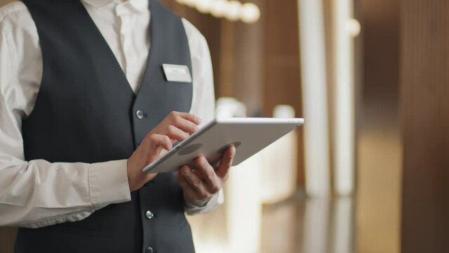 Medium section of unrecognizable hotel worker wearing uniform standing in lobby or lounge using digital tablet