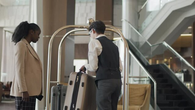 Young Asian bellhop taking African American guests suitcase to bring it to hotel room using luggage trolley