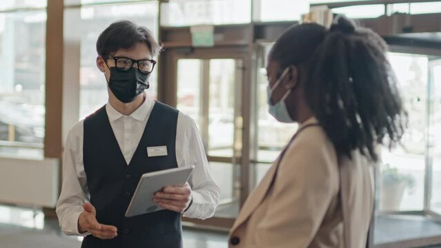 Asian concierge wearing protective mask having conversation with African American guest in hotel lobby