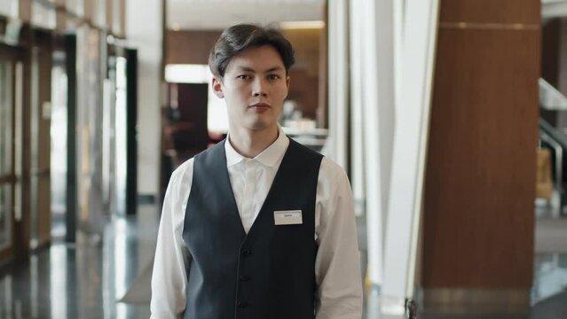 Slow motion medium portrait of young Asian concierge standing in hotel lobby looking at camera
