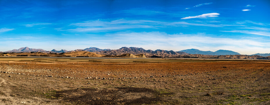 A panorama view ofLake Grassmere farmland with a backdrop of the lake and the rugged mountain range under a blue cloudy sky