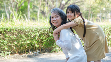 Smiling middle age grandmother giving piggyback ride to cute little granddaughter during walking in the park.