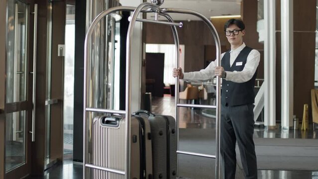 Portrait of modern young Asian hotel porter standing with luggage trolley in lobby looking at camera