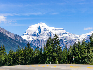 View to Mount Robson with slightly clouded blue sky and highway and green gorest on foreground