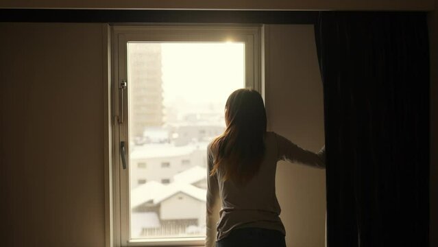Young Asian woman tourist dragging suitcase walking into hotel room. Attractive girl opening curtains and looking cityscape out of the window. Digital nomad work from anywhere and solo travel concept.