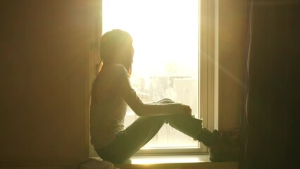 Loneliness Asian woman sitting by the window in bedroom and looking cityscape building covered in snow at winter sunset. Attractive girl resting in hotel room during travel alone on holiday vacation.