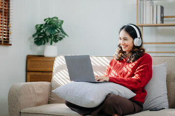 Attractive happy young Asian student studying at the college library, sitting at the desk, using a laptop computer, tablet and headphones having a video chat. in sofa