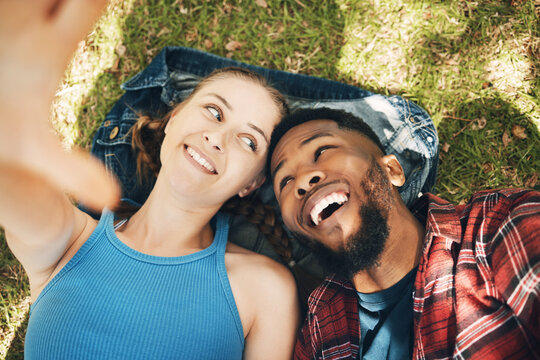Interracial couple, park selfie and lying on grass with smile, love and happiness to relax on holiday. Black man, woman and profile picture on lawn for social media app with romance, above and care