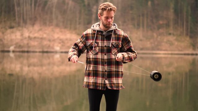 A young man standing next to a beautiful background, where the water mirrors the forest behind. He is playing around with his diabolo and make nice moves. His face is very concentrated and focused.