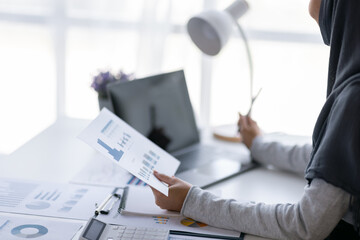 Business Asian muslim woman using calculator and writing make note with calculate doing math finance on an office desk. Woman working at office with laptop and tax, accounting, documents on desk