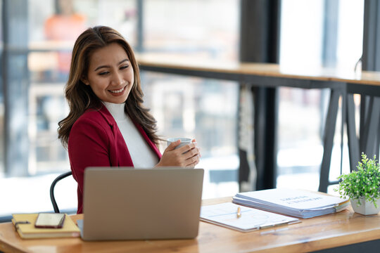 Beautiful asian businesswoman sitting working with laptop computer on table, holding cup of coffee, taking break, relaxing after working on finances. and marketing planning in the office. - Powered by Adobe
