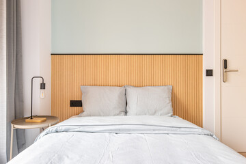 Close up of bed with headboard against wall with wooden decor. Bed is linen in pastel gray. Large window with dark gray curtains. Bedside table with night lamp.
