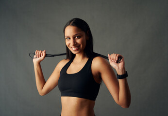 Portrait of happy young biracial woman in sports bra holding jump rope behind neck in studio - obrazy, fototapety, plakaty