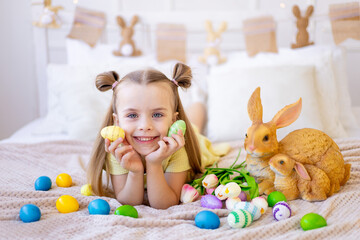 easter, a little girl with painted colored eggs, tulips and a rabbit at home in a bright room is preparing for the holiday smiling, having fun and playing with eggs closing her eyes