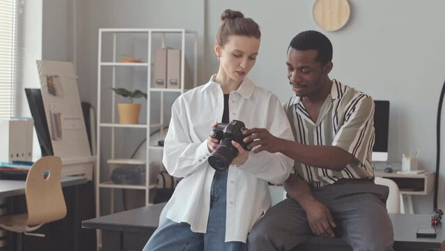 Multiethnic couple of professional photographers discussing photos on digital camera display, working together in bright contemporary photo studio