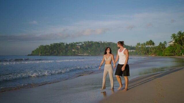 Young biracial happy couple holding hands and walking on the beach together enjoying summer. Cheerful boyfriend and girlfriend relaxing and taking a walk at the seaside hugging and kissing at sunrise.
