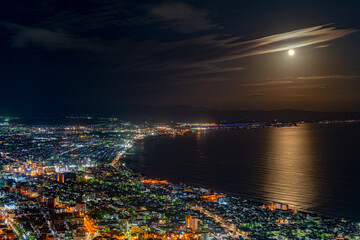 Hakodate City night view from Mt. Hakodate observatory, big bright moon light up the sea, golden reflection on surface. Famous scenic spot in the world. Hokkaido, Japan