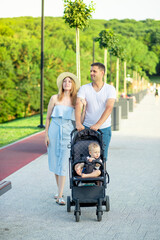 Fototapeta na wymiar happy young parents mom and dad are walking with a baby in a stroller in the park in a hat in the summer in the setting sun and smiling having fun