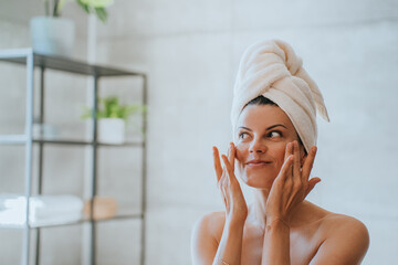 Young adult hispanic woman at bathroom with towel wrapped around head applying cream on face...