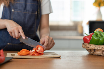 Beautiful Asian woman in kitchen cooking apron preparing various vegetable ingredients and slicing tomatoes in online cooking prep for the health and happiness of our loved ones in their own homes.