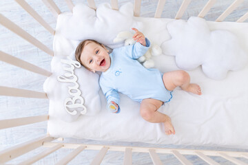 happy smiling baby boy in the crib with the inscription baby in a blue bodysuit, cute joyful little...
