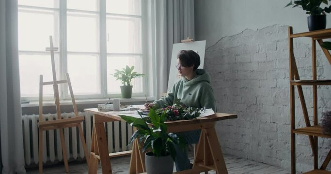 Young woman artist dressed in a hoodie draws pictures on paper with watercolors while sitting at a table in a workshop