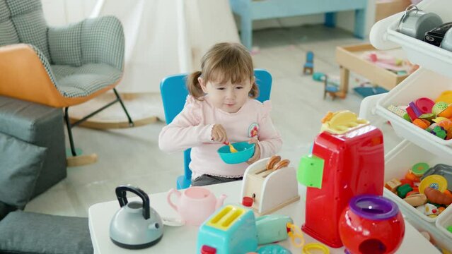 Funny Little Girl Pretend Eating with Toy Spoon From Plastic Empty Bowl  in Kitchen Sitting by the Table in Playroom