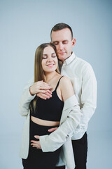 Happy young man standing with his pregnant wife and hugging her belly, isolated on white background. Stylish young people. A man in a shirt and a woman in a black skirt and jacket