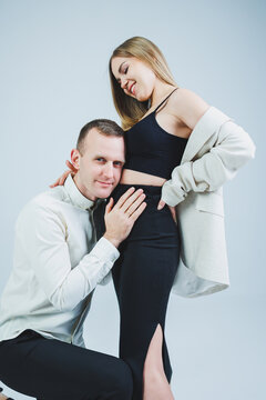 Smiling young man kneeling with his pregnant wife and hugging her belly, isolated on white background. Stylish young people. A man in a shirt and a woman in a black skirt and jacket