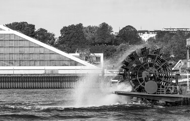 Seitliche Ansicht auf das Rad eines Raddampfers wasserspritzend vor dem teilweise zu sehenden Dockland in Hamburg, schwarz-weiß, horizontal 