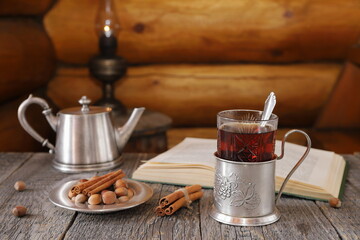 On wooden table on background of old lantern standing on wooden barrel near log wall is glass of tea in iron holder next to iron plate with nuts, iron teapot and book. (Text of book is in English.) 