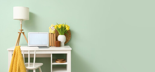Workplace with modern laptop, tulips and Easter rabbit with greeting card near light green wall....