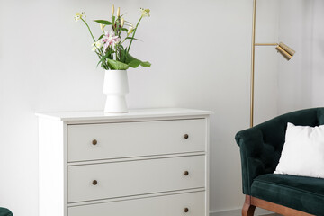Vase with flowers on white chest of drawers near light wall