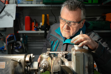 A man in safety glasses and work uniform sets up a grinding machine in his workshop. The grinding machine details in focus. In the background are various working tools on metal shelving for workshop