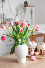 Vase with tulips, Easter eggs and cakes on counter in kitchen