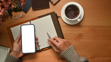 Above view of woman hands holding smart phone and making notes on notebook over wooden desk