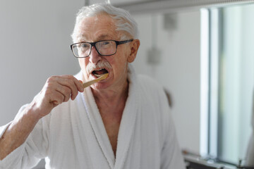 Senior man cleaning his teeth with wooden brush in bathroom, sustainable lifestyle.