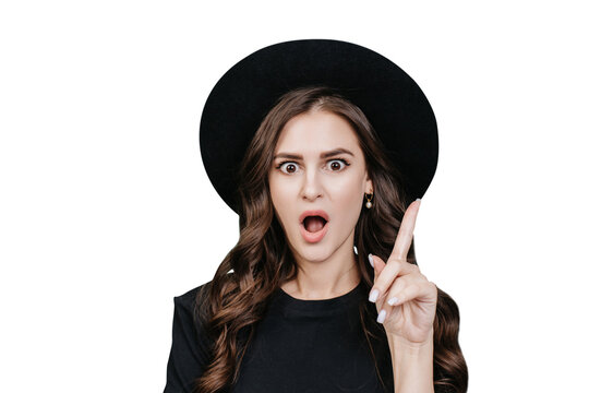 Great idea! Attractive hispanic young woman in black hat and black t-shirt with wide open eyes and mouth raises index finger with surprised face against transparent background. Look up, that's amazing