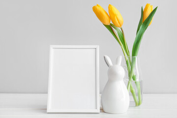 Blank frame, Easter bunny and vase with yellow tulip flowers on white table near grey wall