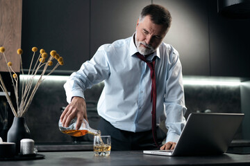 Upset senior man drinker alcoholic standing in kitchen holding glass drinking whiskey alone