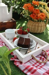 a piece of chocolate cake, a bowl of strawberries and a cup of tea on a wooden tray against a background of flowers in a vase