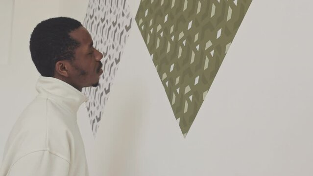 African American man standing close to minimalist pattern pictures on white wall in modern art gallery examining them