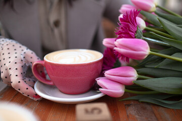 Fototapeta na wymiar Close---up of female hands in elegant stylish pea gloves hold a mug of a coffee. 35 years old woman is drinking coffee or tea in cafe with a bouquet of flowers in the spring streets of the city. 