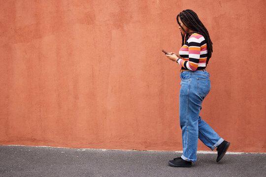Black woman walking with smartphone in city, outdoor with technology, chat and communication with travel and fashion. Social media, urban street and wall with mockup and 5g, female in Jamaica