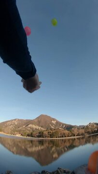 hands juggling 4 balls (green, pink, yellow, orange) in front of Veynes lake reflecting the mountain in background. rocks in bottom foreground. immerssive vertical footage: wide, mid low angle view