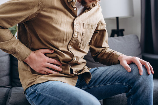 Sad young man sitting on couch at home, suddenly feeling strong stomach ache, gastritis problem. Upset male 30s squeezing belly with hand because of abdominal pain. Guy suffering from stomach ache